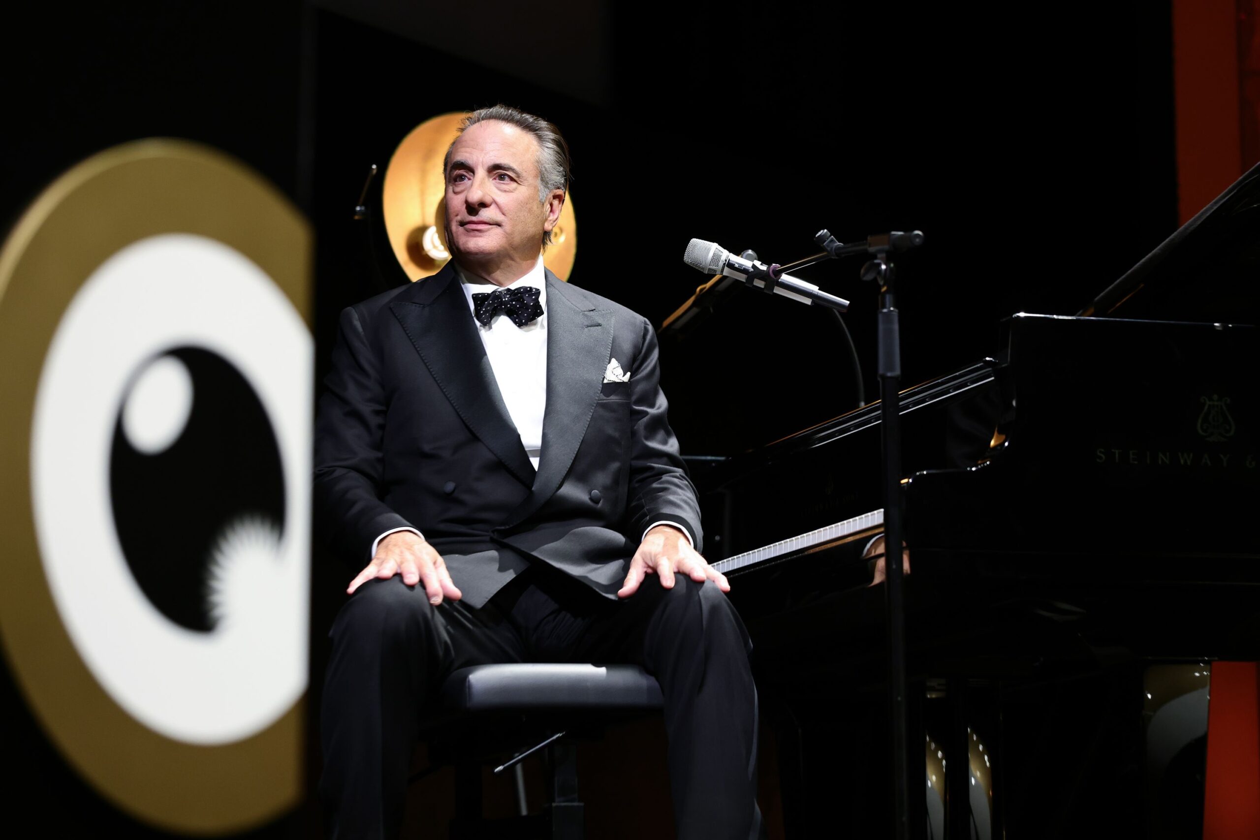 This image shows a man dressed in a black tuxedo with a black bow tie, sitting on a stool in front of a grand piano. The piano is a Steinway & Sons model, as indicated by the logo on the piano. The man appears to be performing or preparing to perform, as he is positioned near a microphone on a stand. Behind him, there is a large circular logo with…